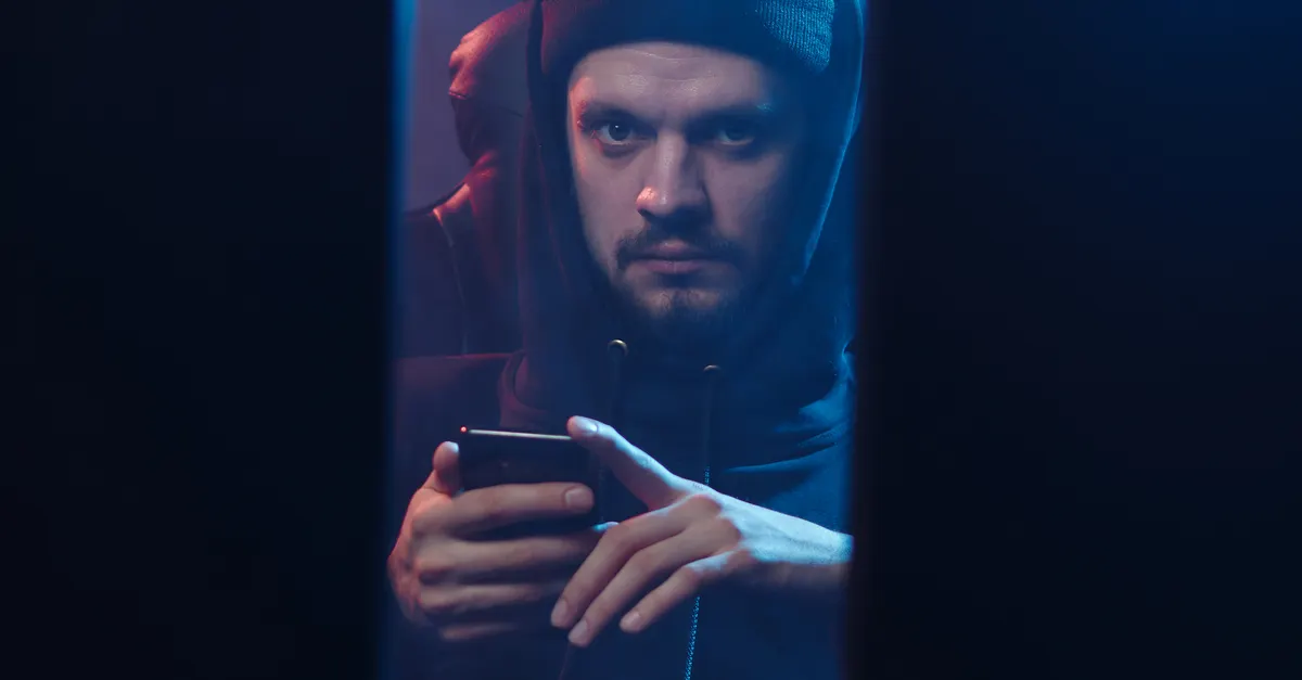 Hooded hacker holding a smartphone in a dark room, illustrating voice phishing attacks