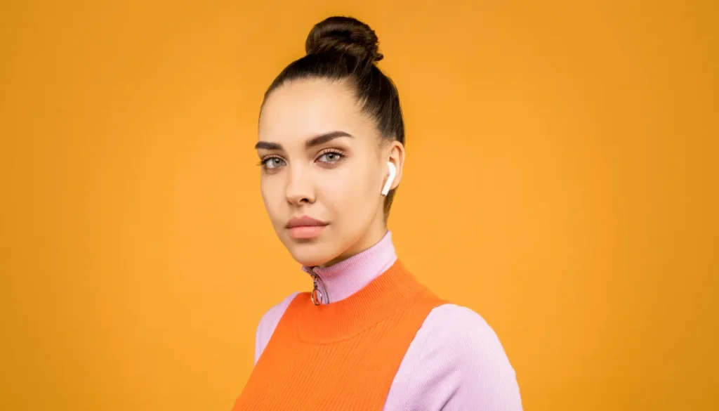 Portrait of a young woman wearing wireless earbuds against a vibrant orange background