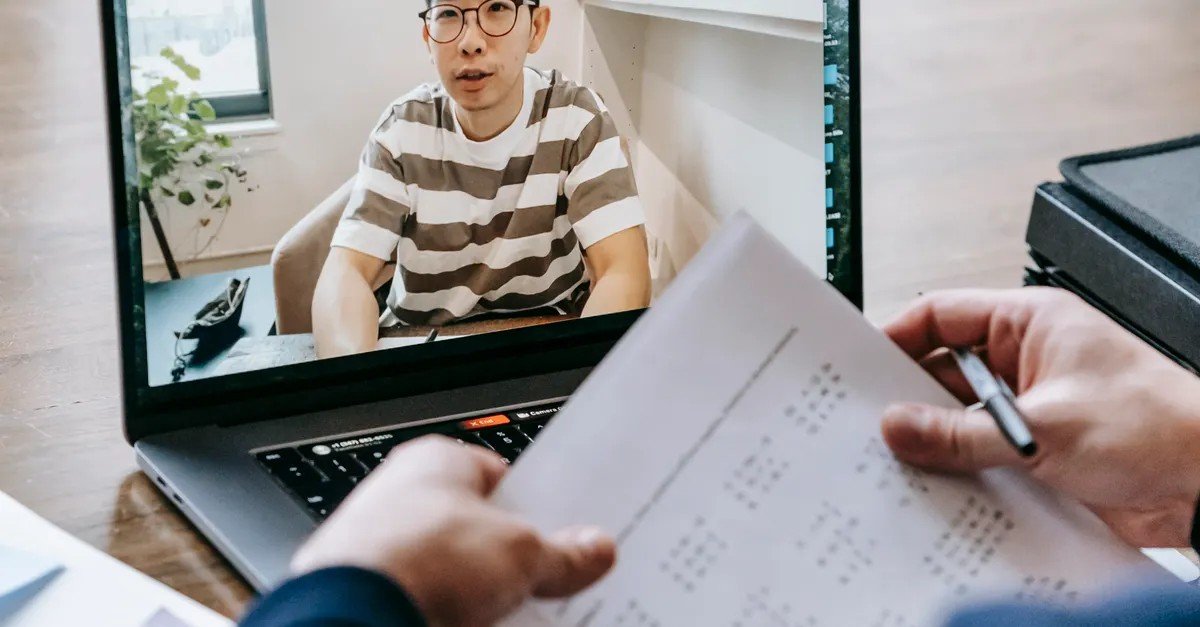 A person appearing on a laptop screen during a video call, while someone holds interview papers in the foreground