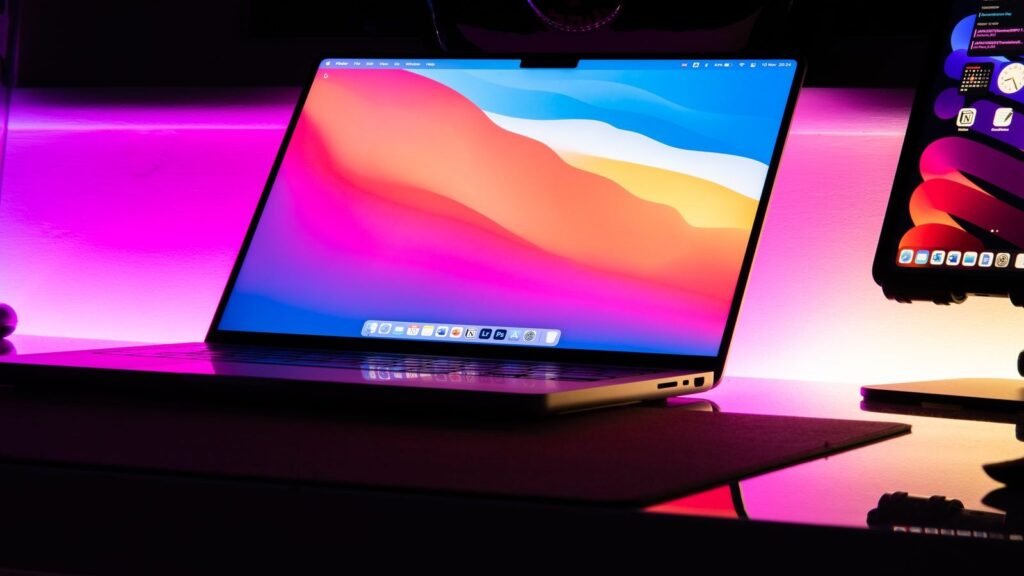 A MacBook glowing with pink and blue neon light on a dark desk