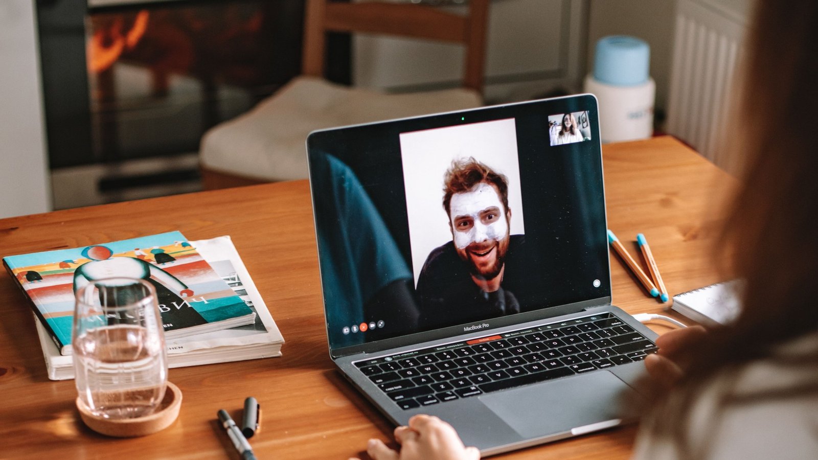 Woman on a video call on a MacBook Pro at a wooden desk with a notebook and glass of water