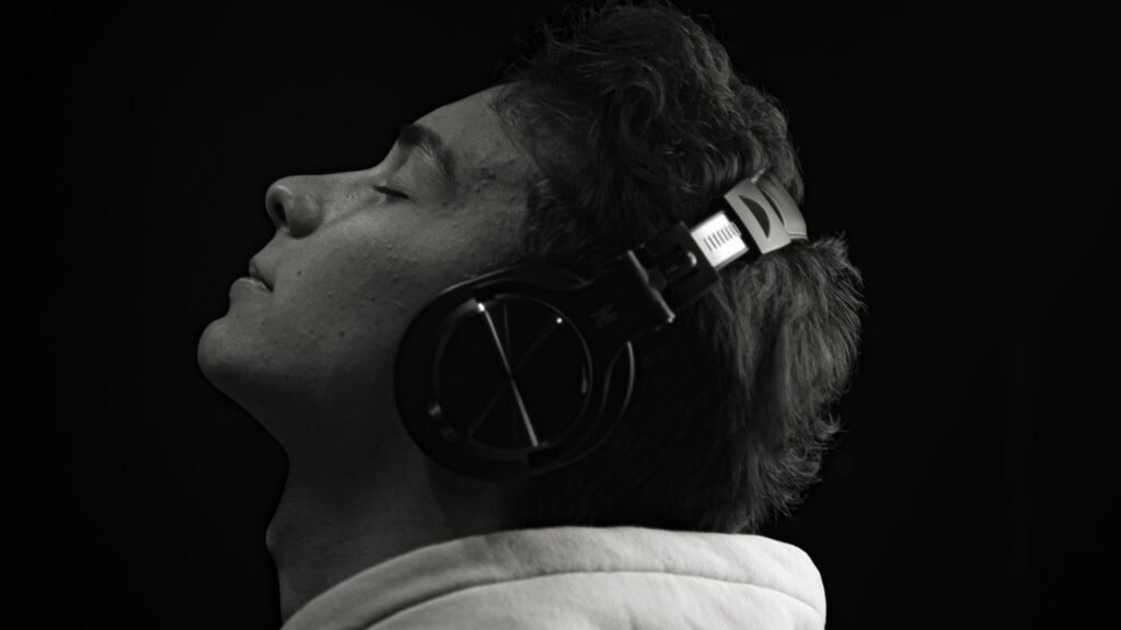 Young man with eyes closed wearing over-ear headphones, head tilted up against a black background