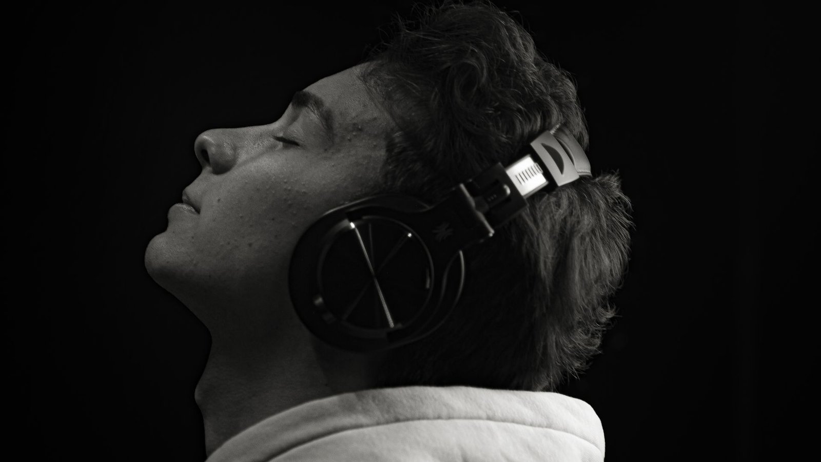 Young man with eyes closed wearing over-ear headphones, head tilted up against a black background