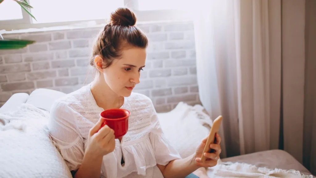 Woman holding a red coffee cup and reading her phone in bright morning light