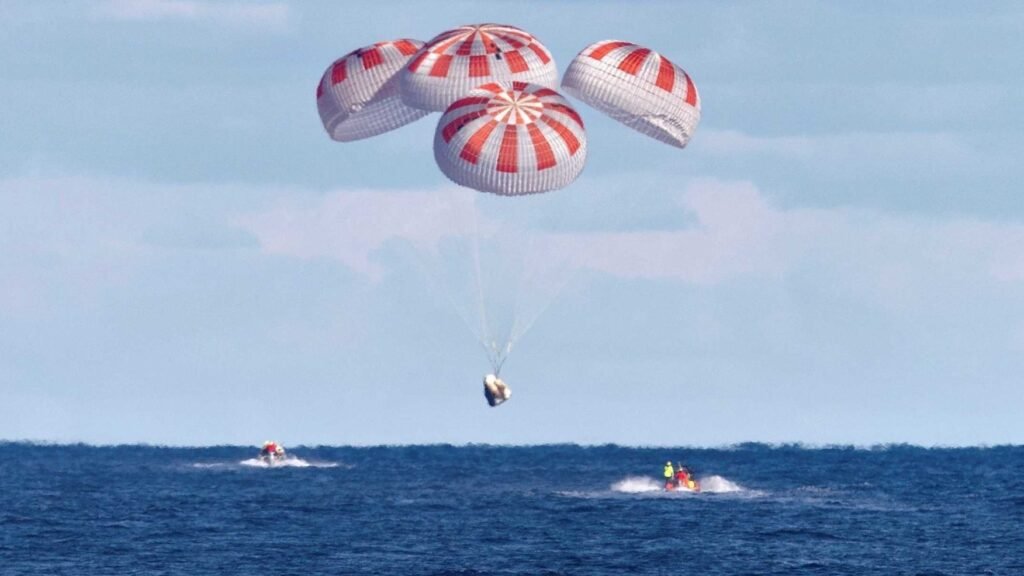 A SpaceX Crew Dragon capsule descends under four red-and-white parachutes toward the ocean as recovery boats approach.