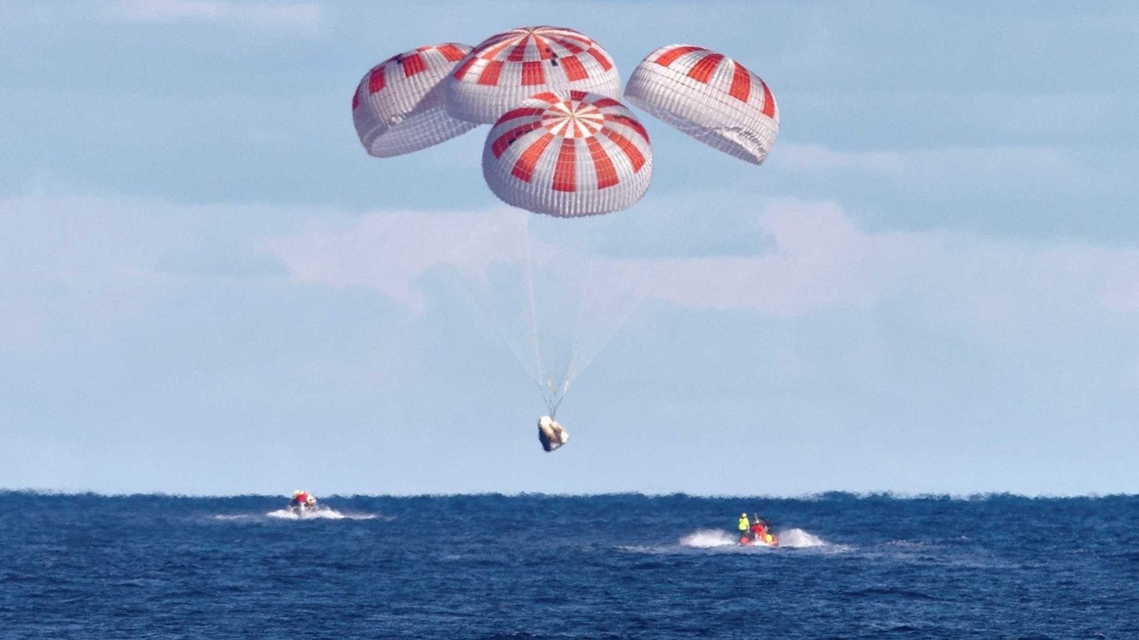 A SpaceX Crew Dragon capsule descends under four red-and-white parachutes toward the ocean as recovery boats approach.