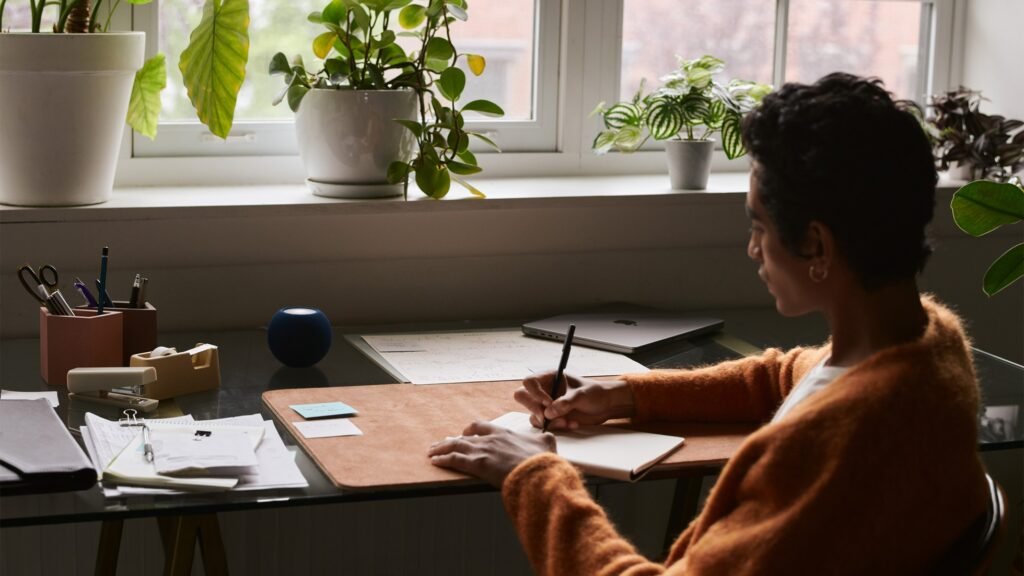 A person working at a desk with an Apple HomePod mini smart speaker and a MacBook