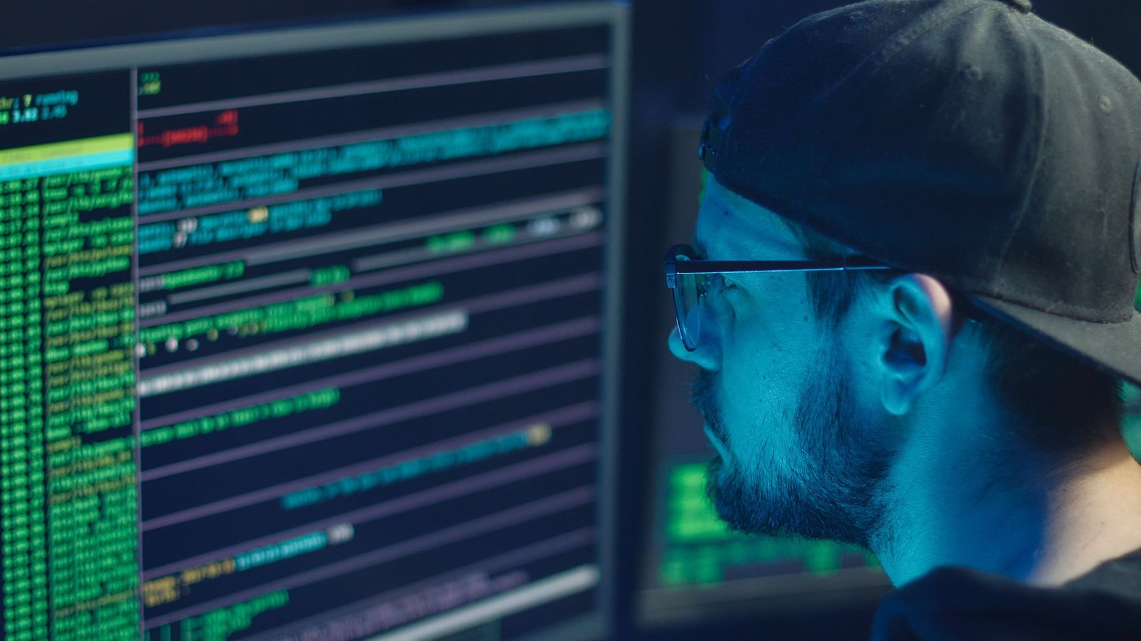 A cybersecurity analyst inspecting code on monitors in a dark office, lit by blue screen glow