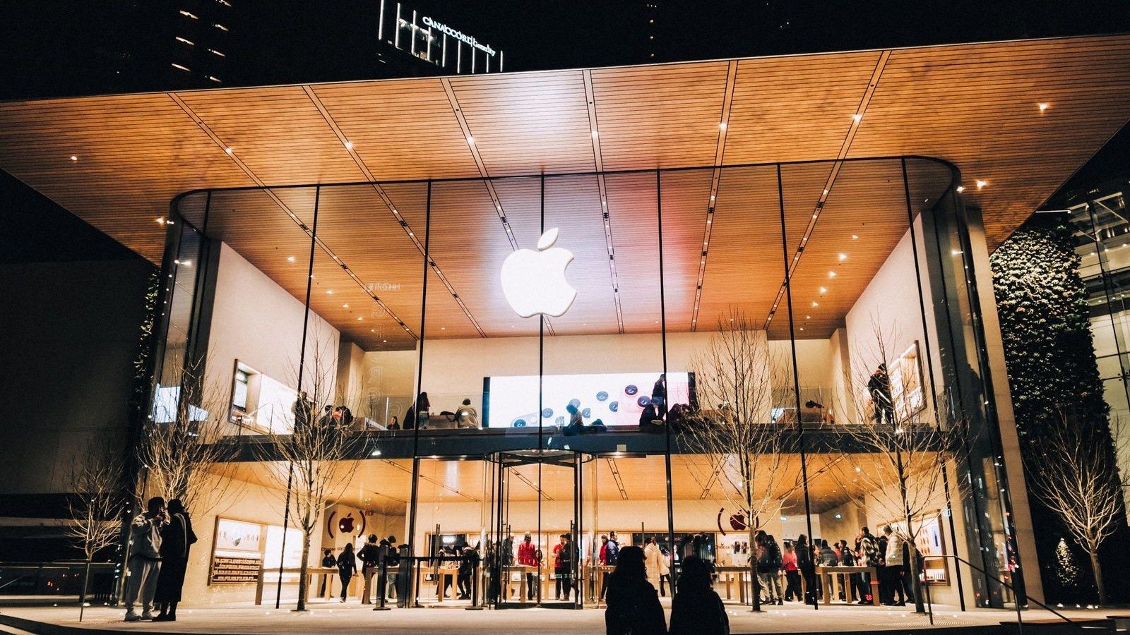 Crowds inside a glowing Apple Store at night beneath a lit Apple logo