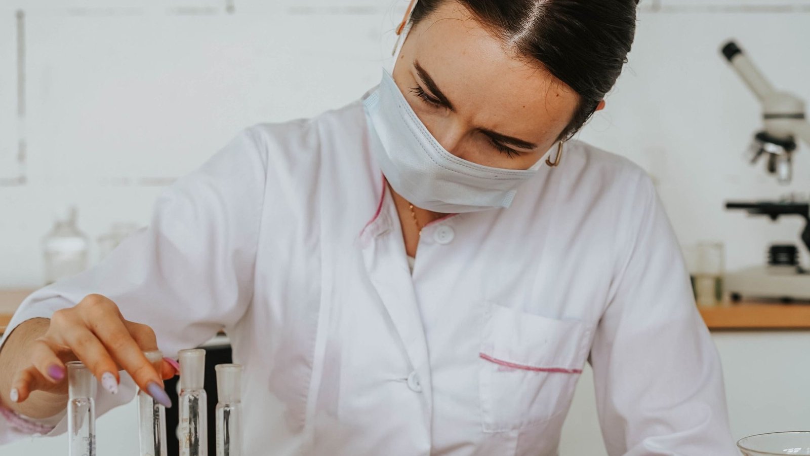Scientist in a white coat and face mask handling test tubes in a pharmaceutical research laboratory