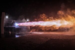 Rocket engine firing at night, sending a bright plume of flame and smoke across a test site