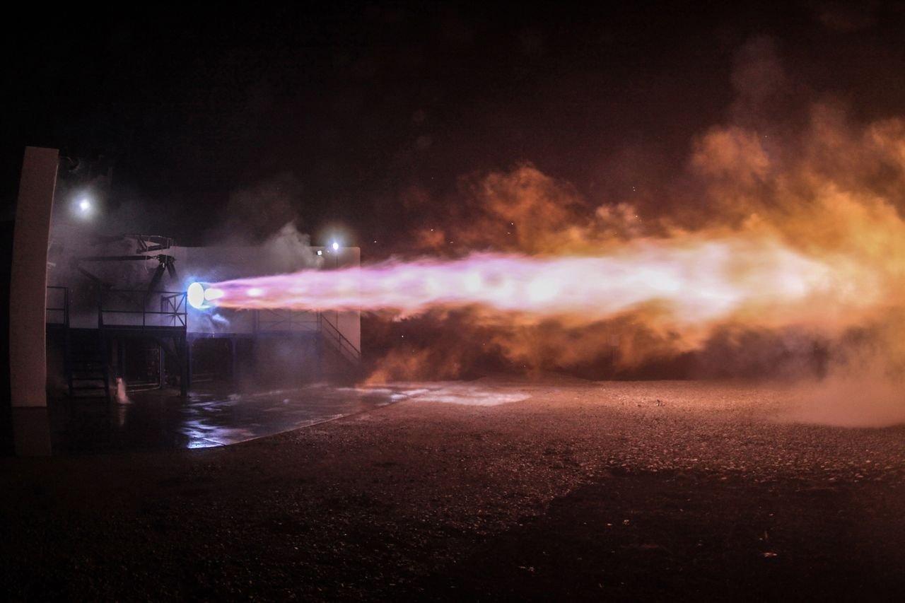 Rocket engine firing at night, sending a bright plume of flame and smoke across a test site