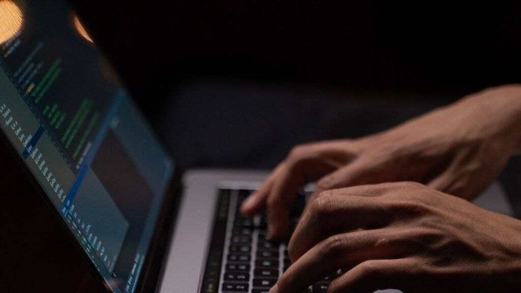 Hands typing on a laptop keyboard with code on screen in a dark room