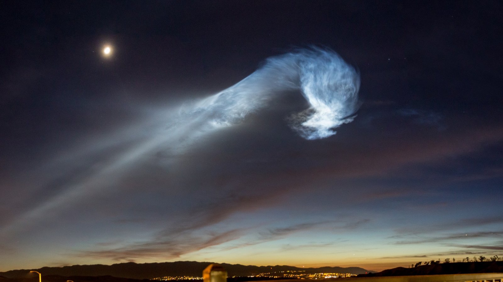 Glowing exhaust plume from a SpaceX Falcon 9 launch over Vandenberg at night, with the moon in the dark sky