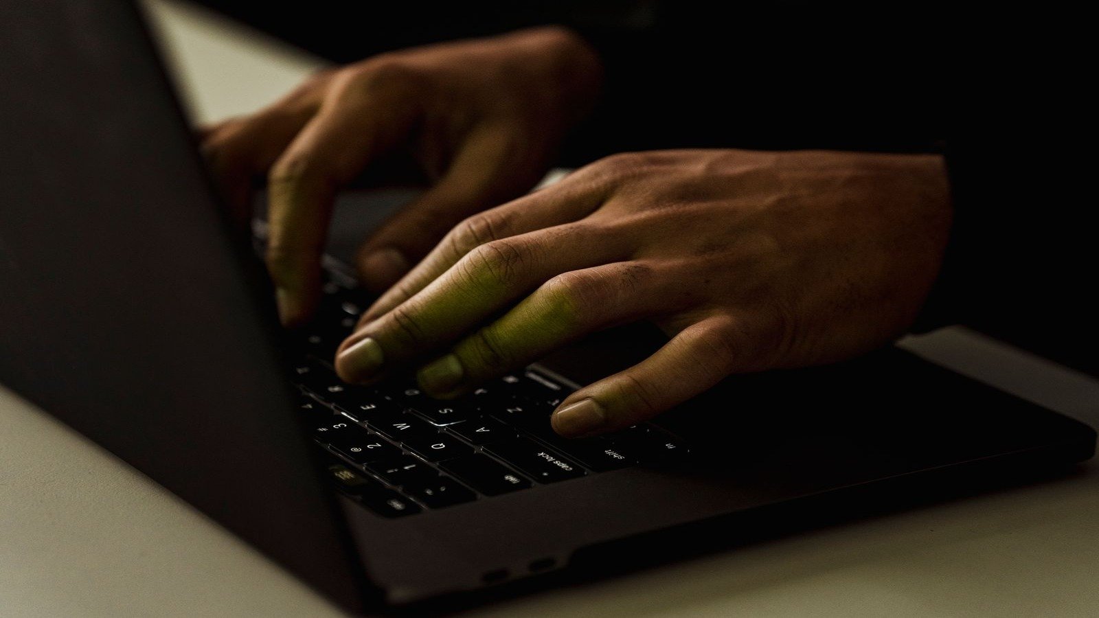 Hands typing on a backlit laptop keyboard in a dark room, the screen's glow falling on the hands.