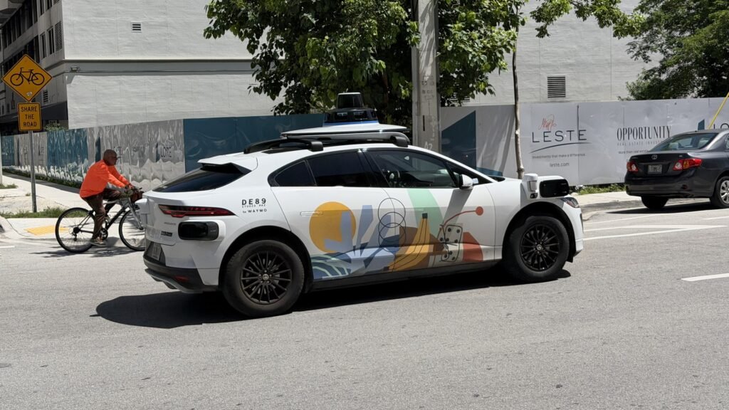 Waymo Jaguar I-PACE robotaxi with rooftop sensor array on a Miami street as a cyclist rides past